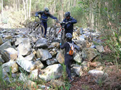 3 ciclistas a carregarem as bicicletas sobre um monte de pedras